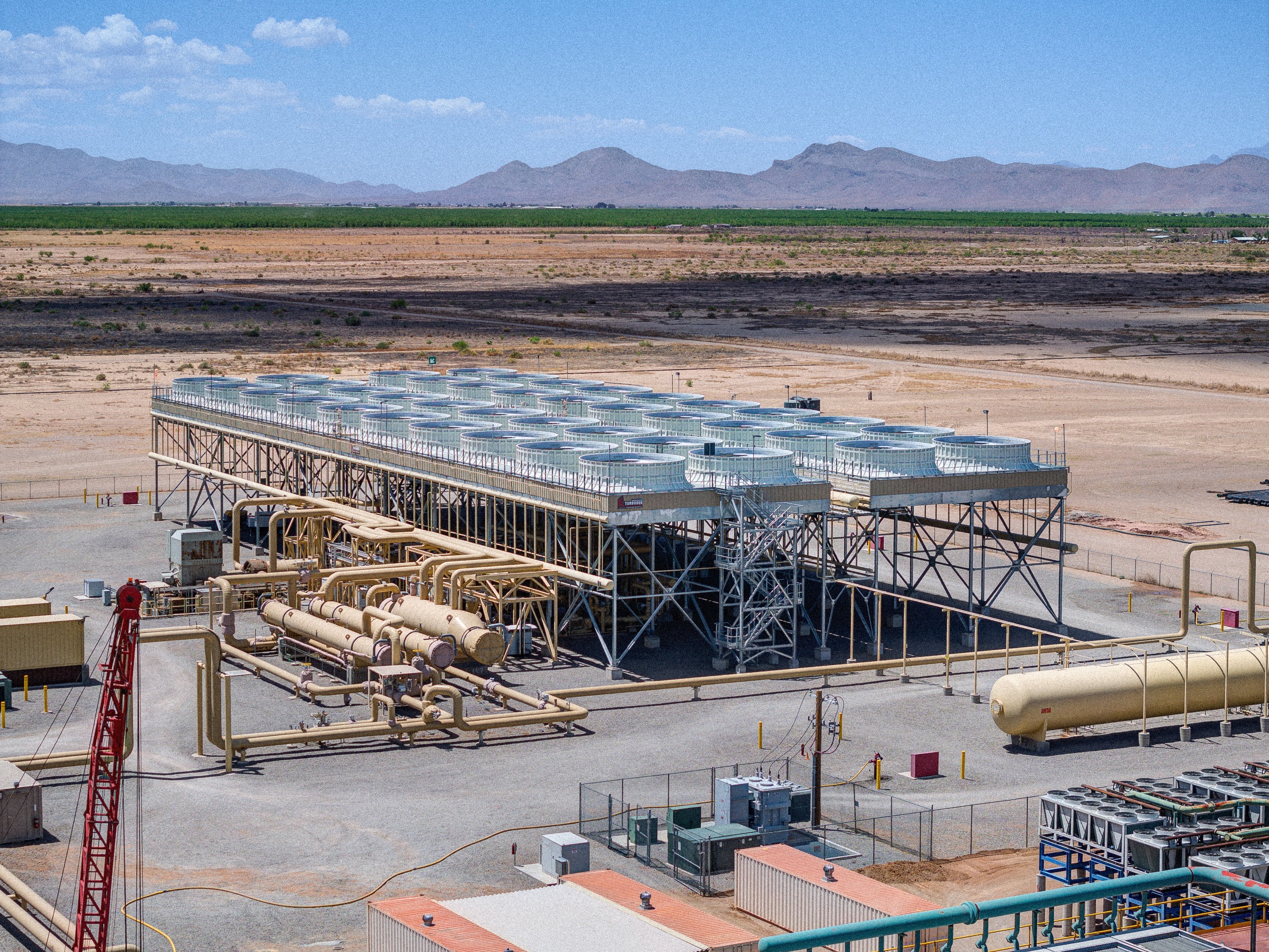 Wide-angle view of the Lightning Dock geothermal power plant in southern New Mexico, showing a long row of silver cooling fans atop a raised steel platform, with yellow steam pipes and equipment in the foreground. The plant sits in an arid desert landscape with fields, sparse vegetation, and distant mountains under a clear blue sky.