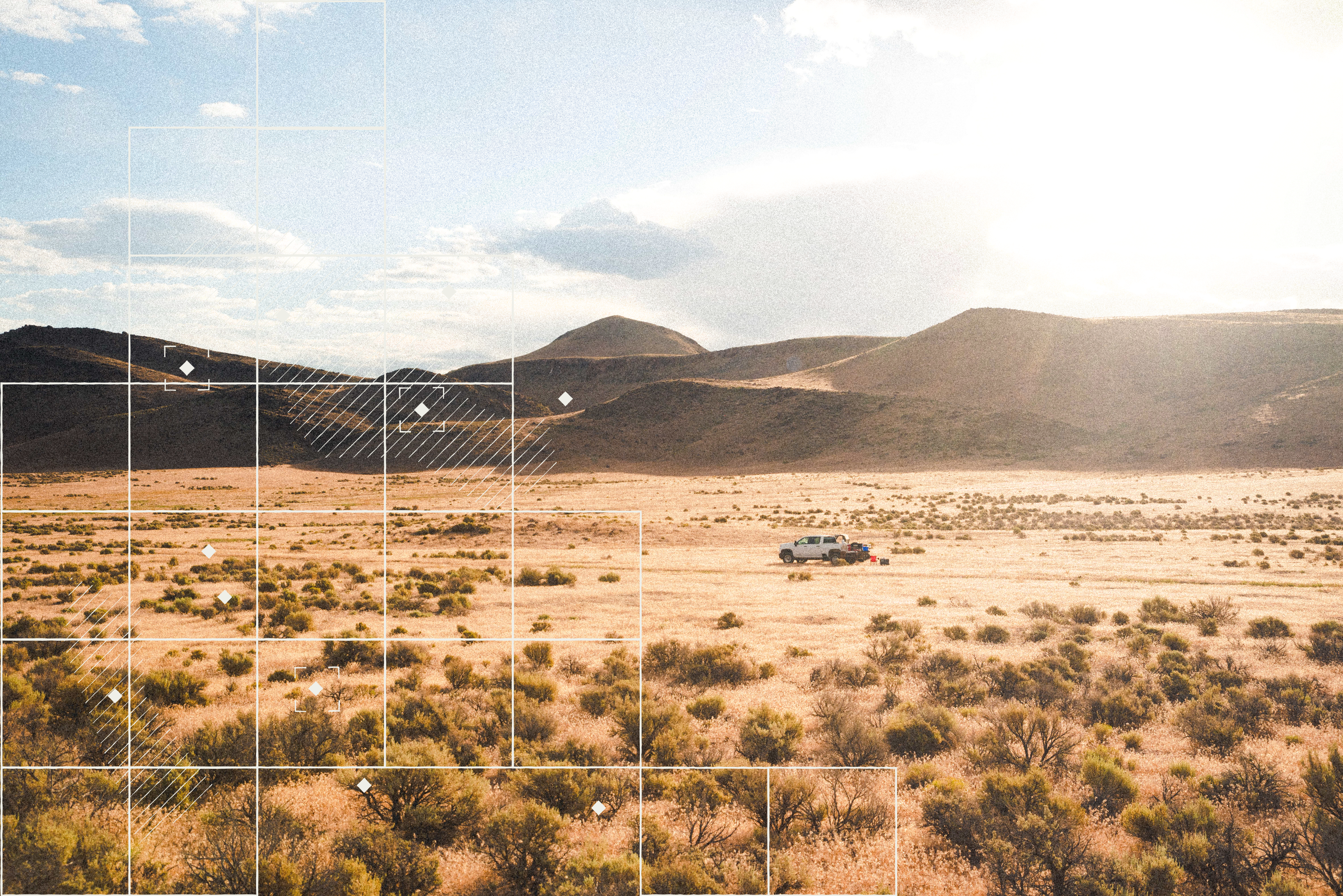 A white field truck sits in a golden desert valley at sunset, surrounded by scattered sagebrush with dark hills rising in the distance. A geometric grid overlay suggests mapping or geological survey work, symbolizing Zanskar’s field-based exploration.
