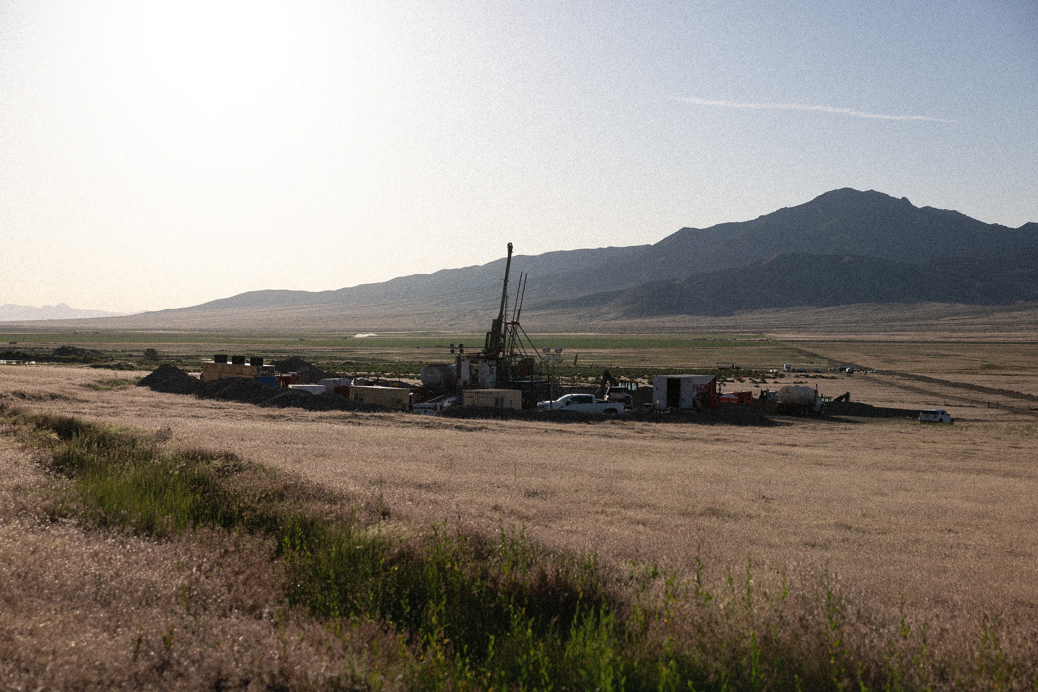 Landscape with dry grassy fields, drilling equipment, and trucks in the middle ground, with mountains in the background. 