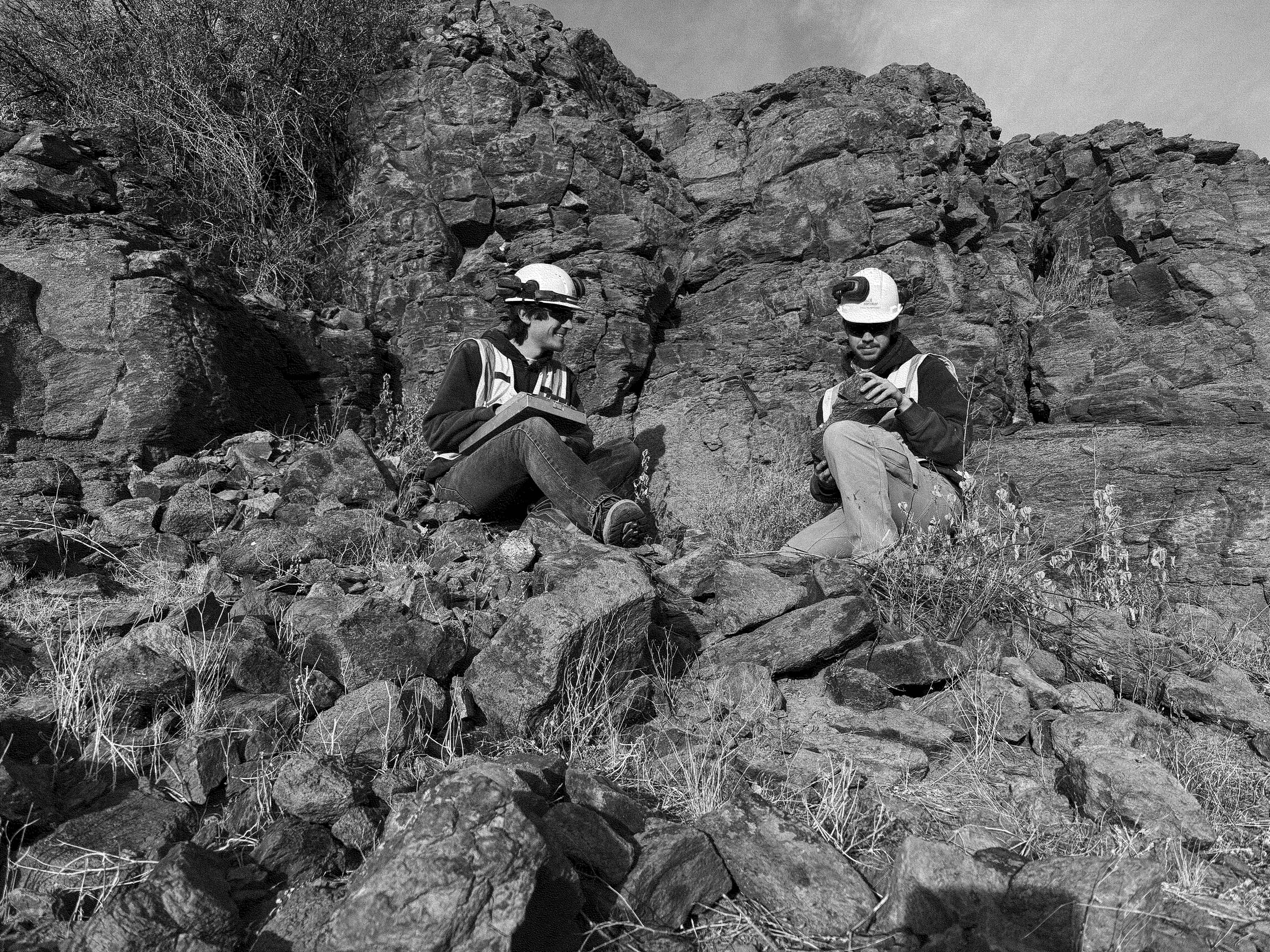 Black and white photo of two geologists wearing helmets and safety vests, sitting on rocky terrain and examining notes and rock samples in front of a rugged cliffside. The image is framed with a Zanskar light teal border.